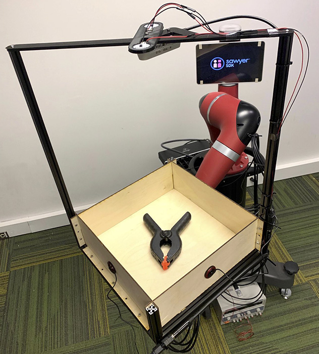 The Tilt Bot has a robotic arm with a wooden tray attached to it. Inside the tray is a metal tool. A camera hangs above the tray.