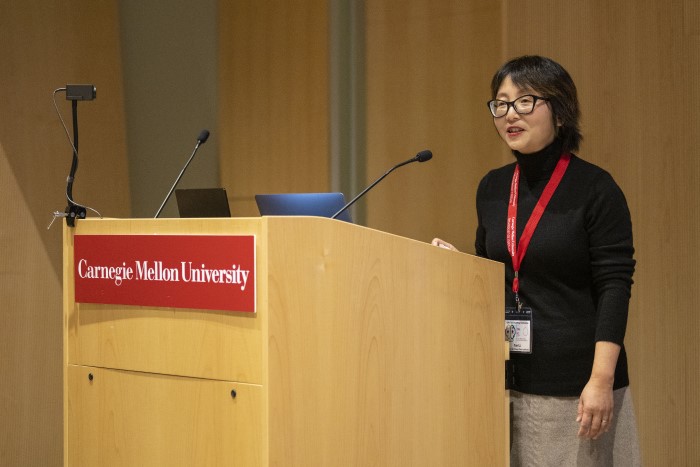 An image of a speaker presentation. The speaker is standing at a podium while explaining what is being depicted on the projector screen.