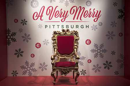 A photo of an ornate red chair infront of a snow flake backdrop with a sign for A Very Merry Pittsburgh above