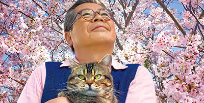 Promo image for the Opening Night with The Island of Cats at the Pittsburgh Japanese Film Festival, man holding a cat in front of cherry blossoms