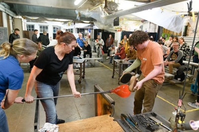 Image of three people working on a glass blowing peice in front of a crowd