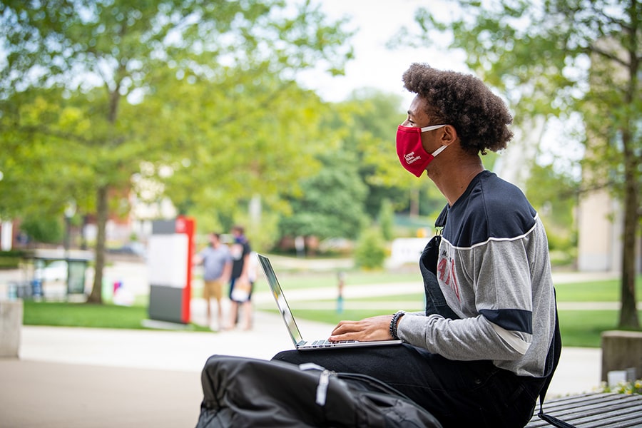 a CMU student wearing a facial covering sitting with his lap top on campus on a nice day