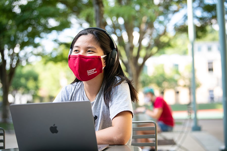 a student in a face covering working on her laptop on a sunny day on campus