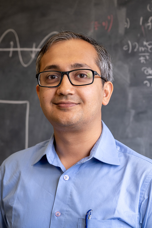Shubhayu Chatterjee smiles for the camera. He is standing in front of a chalkboard and is wearing glasses and a blue button-down shirt.