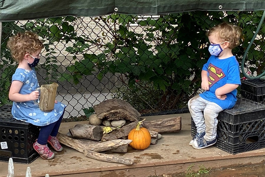 two toddlers sitting outside playing at a distance and wearing facial coverings