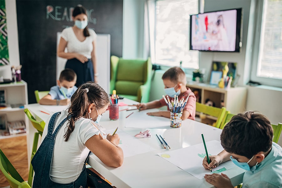 Image of four children wearing masks, socially distanced and working at a table with teacher in background