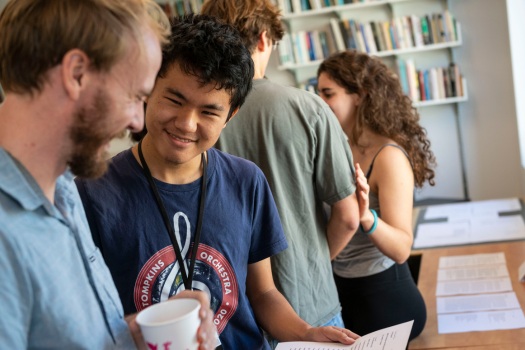 A professor and student consult a document. In the background, two other students stand in front of a wall of books.