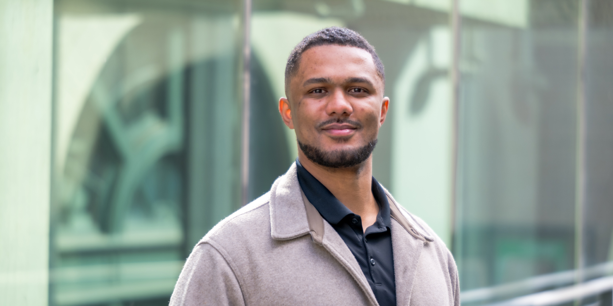 Portrait of a young man with brown hair, short beard wearing a black polo and a beige jacket.