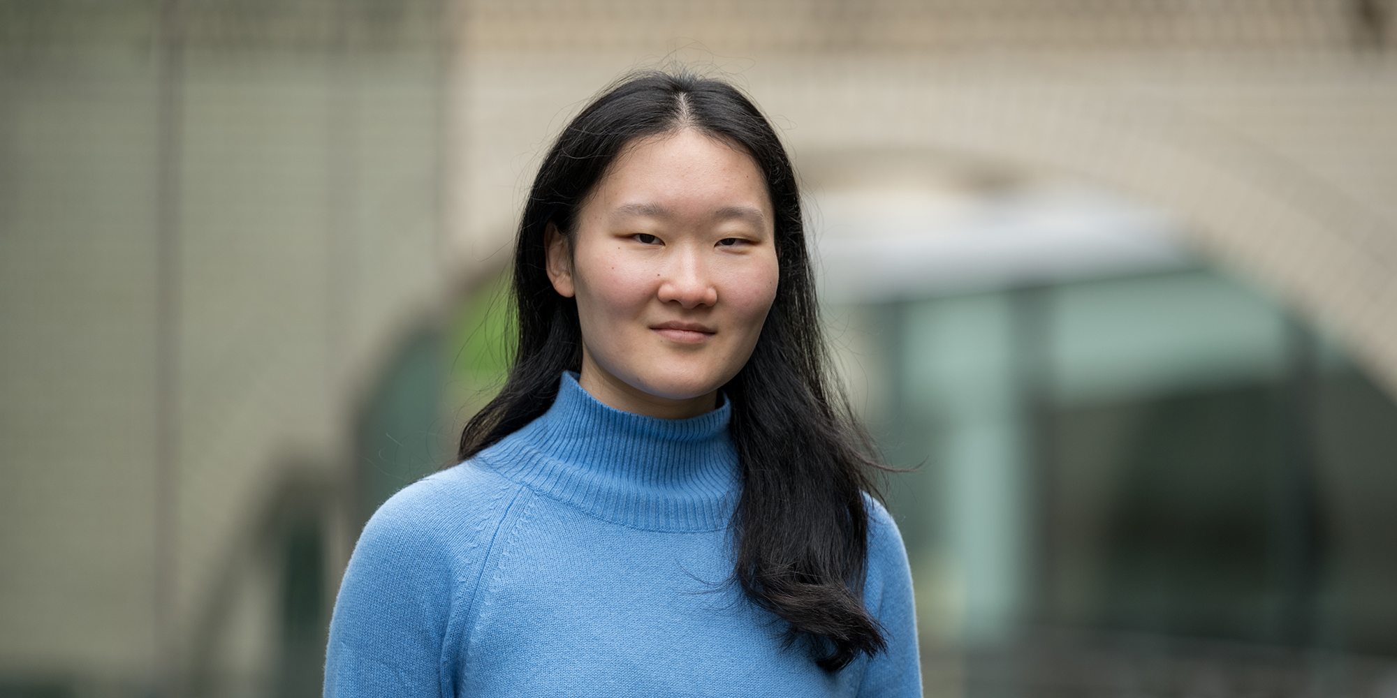 Yunshu Li stands outside of a glass building and a brick archway, and she smiles at the camera.