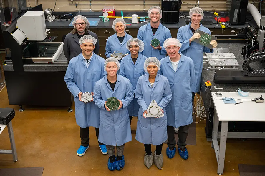 Nine people wearing hairnets and lab jackets and holding components of a module that will be installed into a particle detector.
