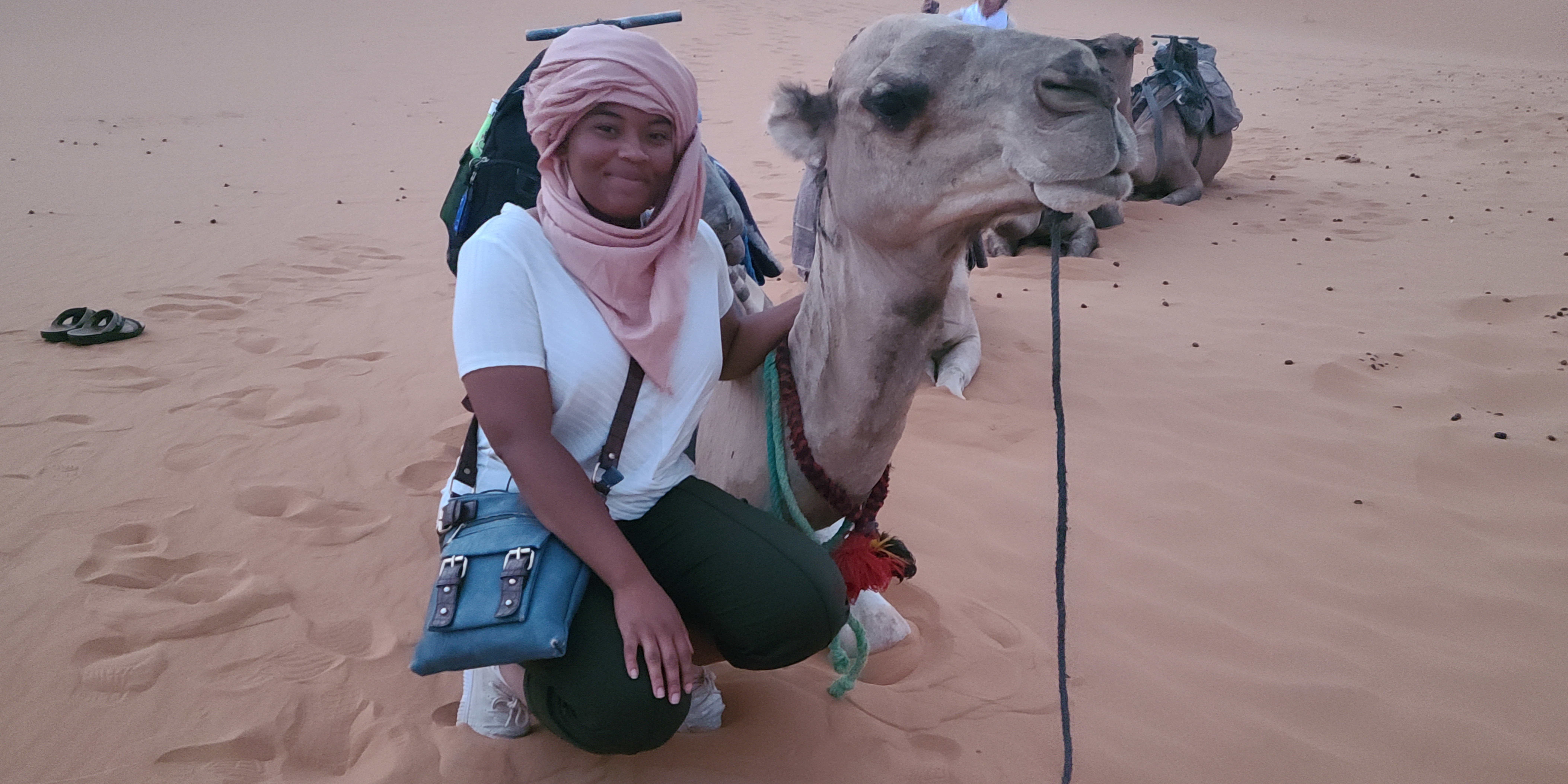 Young student wearing a turban, standing in a desert beside a camel resting on the sand, posing calmly against the backdrop of dunes.