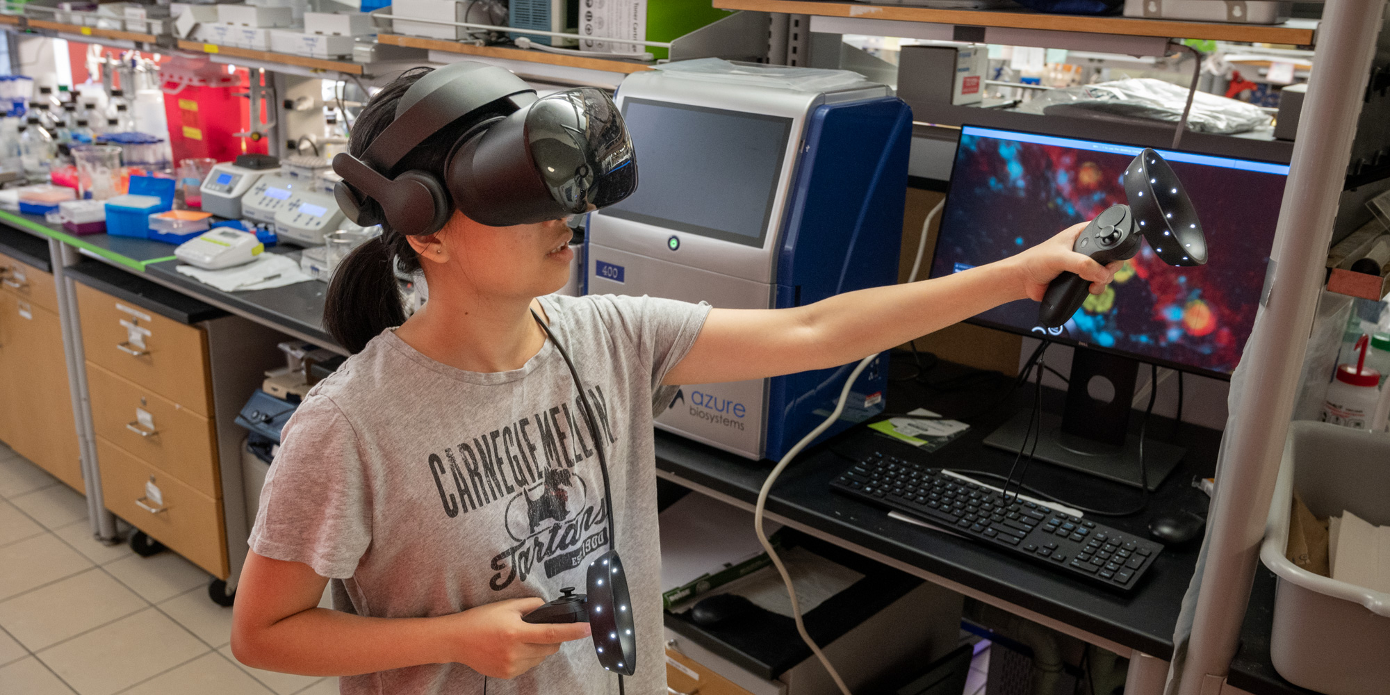 A woman wearing a virtual reality headset moves her arms while a monitor shows the interior of an infected cell that she is navigating.