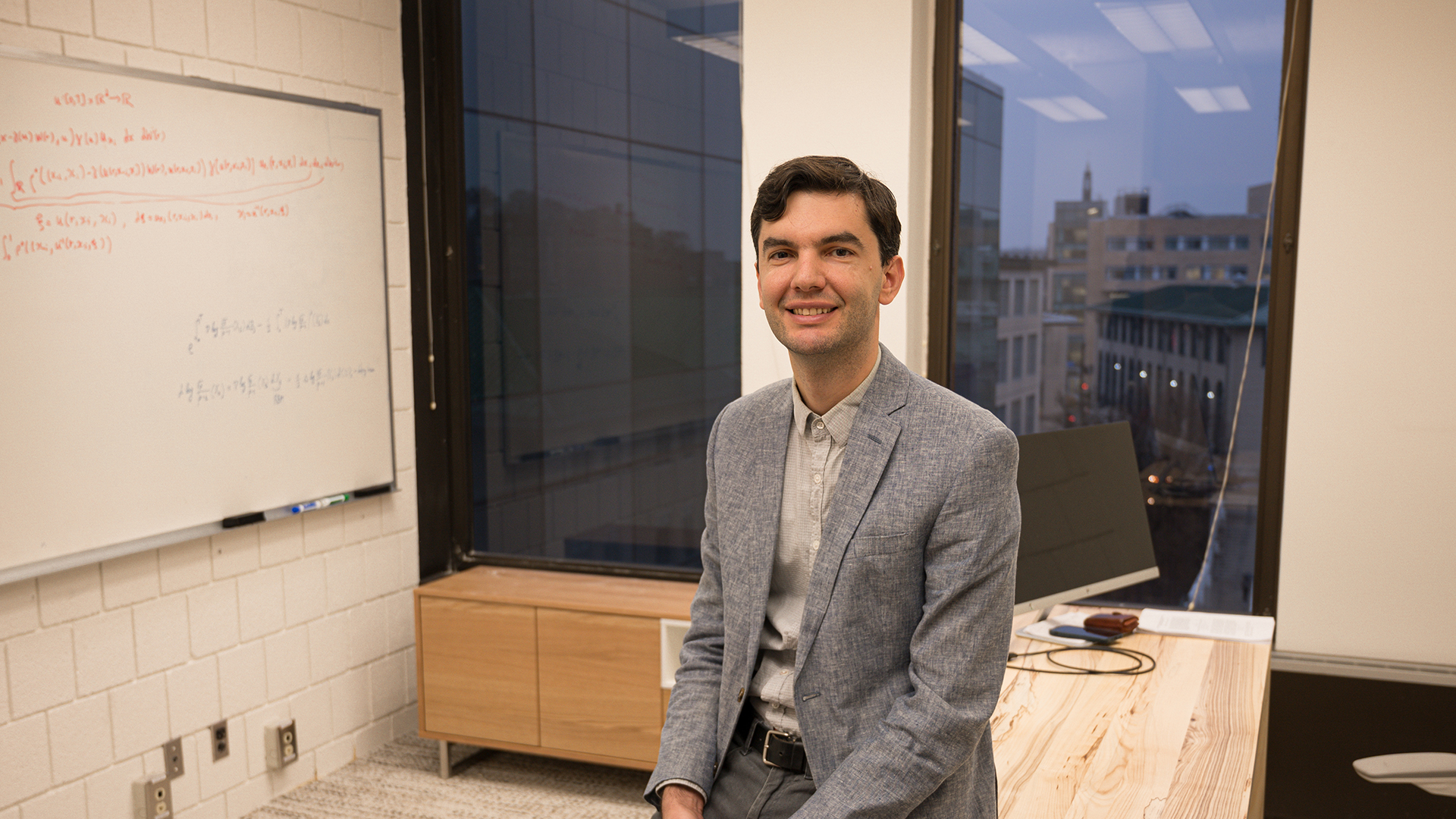 Mykhaylo Shkolnikov leans against his desk in his office and smiles at the camera.