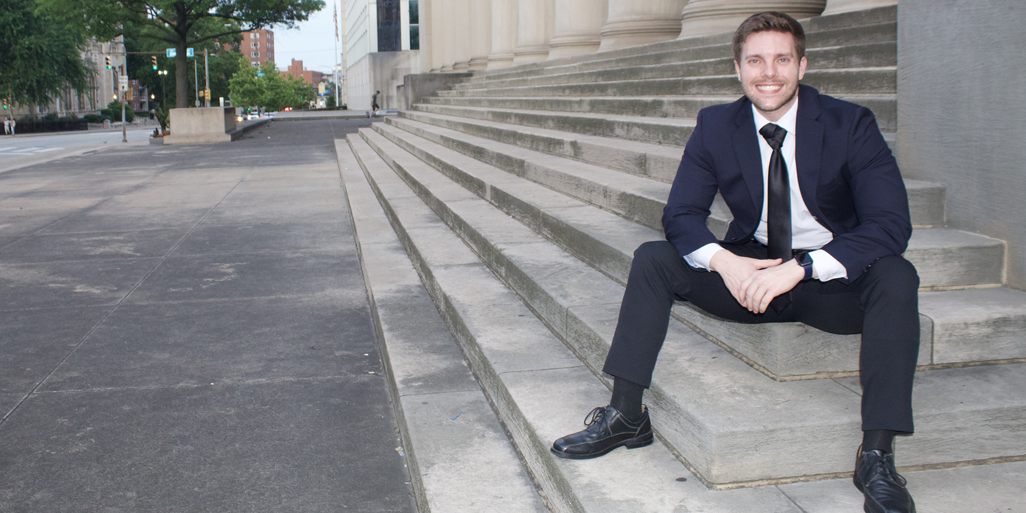 Zach Baldwin in business attire sitting on the entrance stairs of a building
