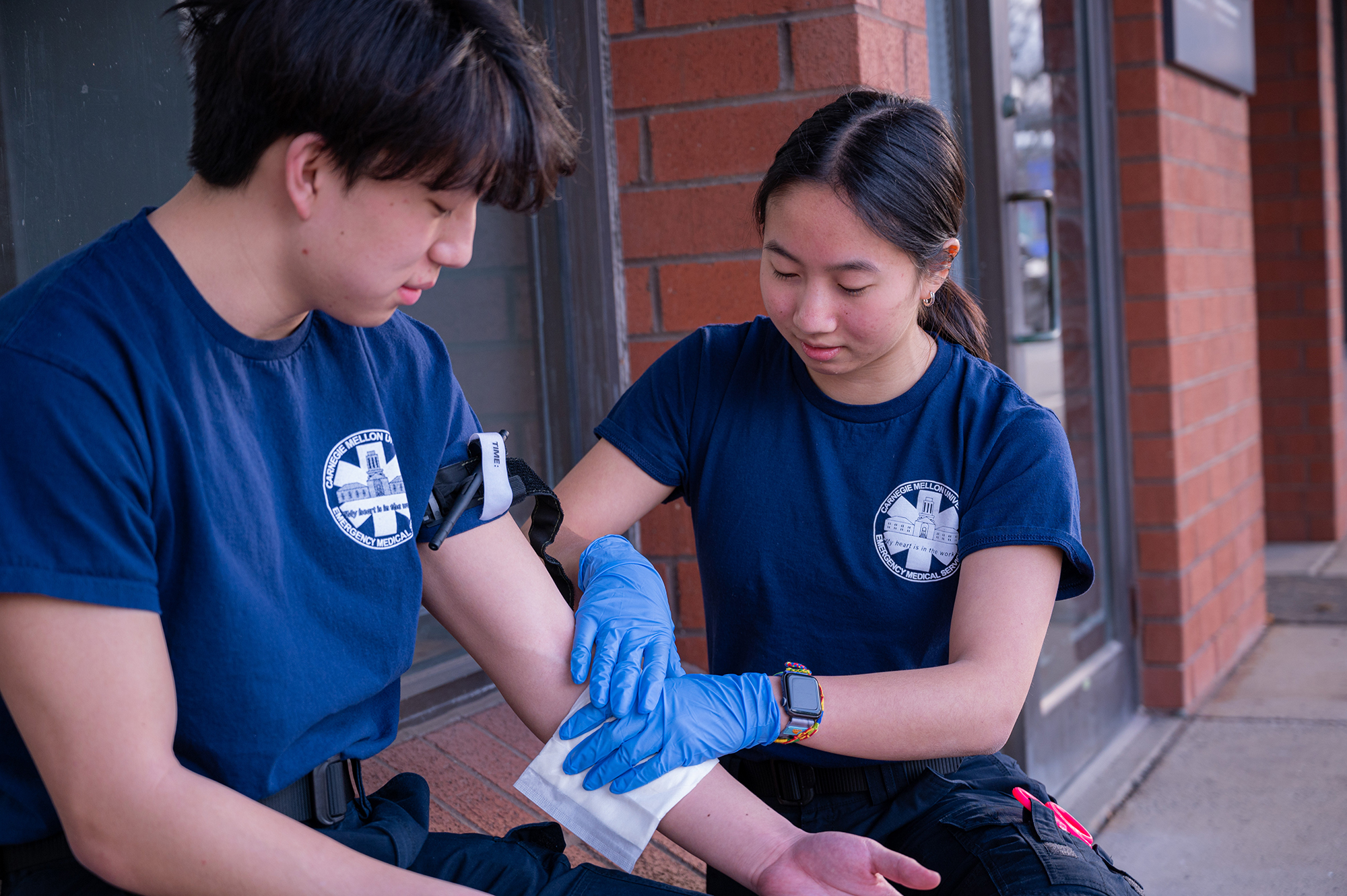 Young female student wearing blue latex gloves attentively practices bandaging a simulated wound on the forearm of a seated male student during a first aid training session.