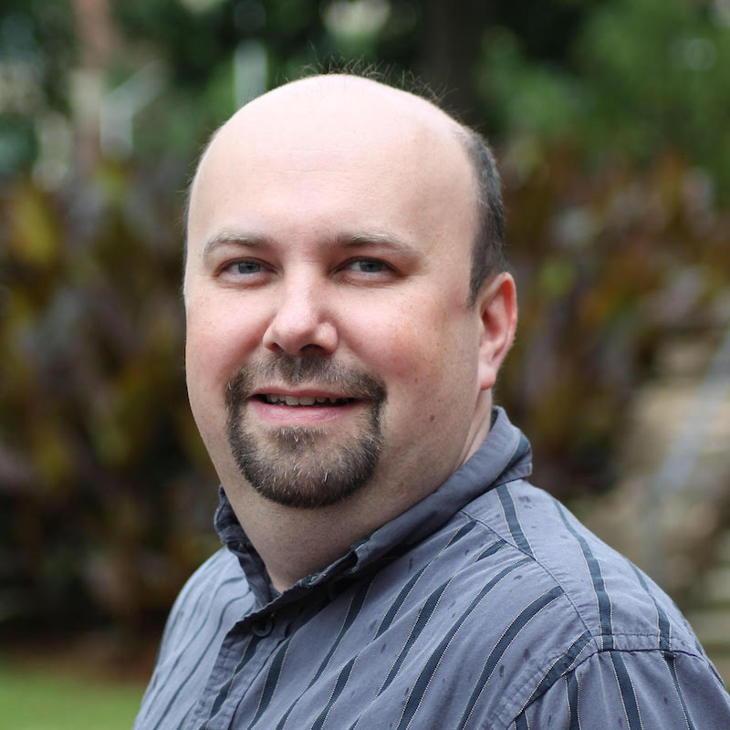 Bald man with a short beard wearing a striped blue-gray shirt, smiling subtly while facing the camera.
