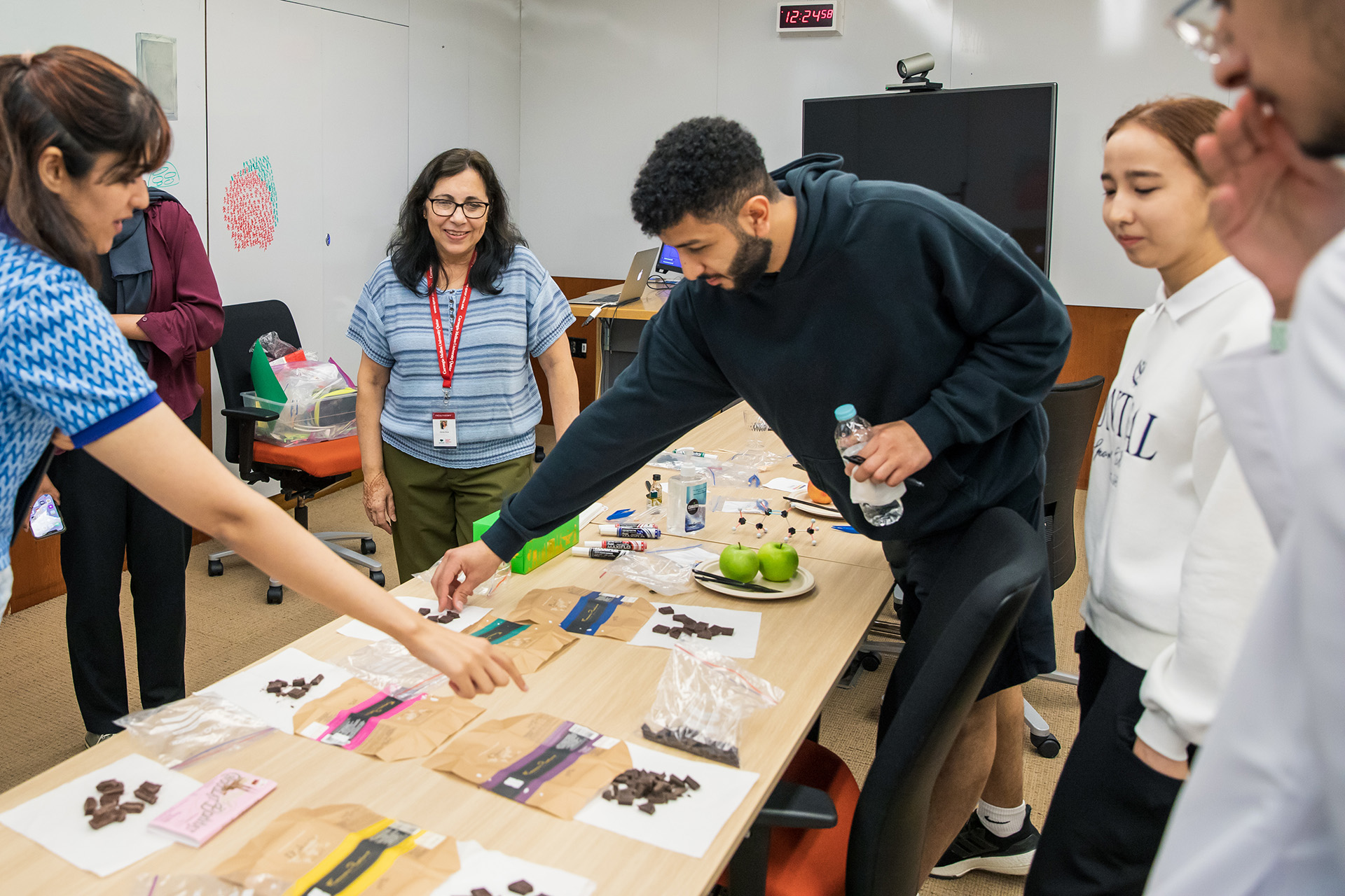 Six individuals gathered around a rectangular table during a chemistry microcourse, with two participants selecting pieces of chocolate as part of a hands-on activity.