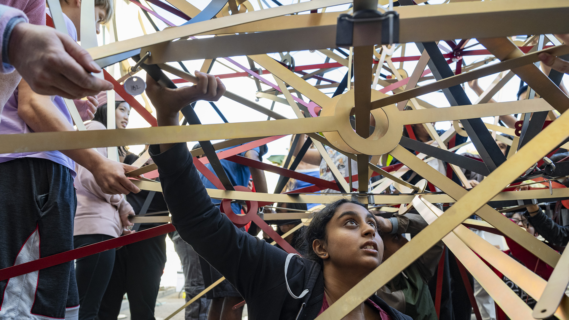 A student holds up a sculpture made of gold, red and black strips of metal.