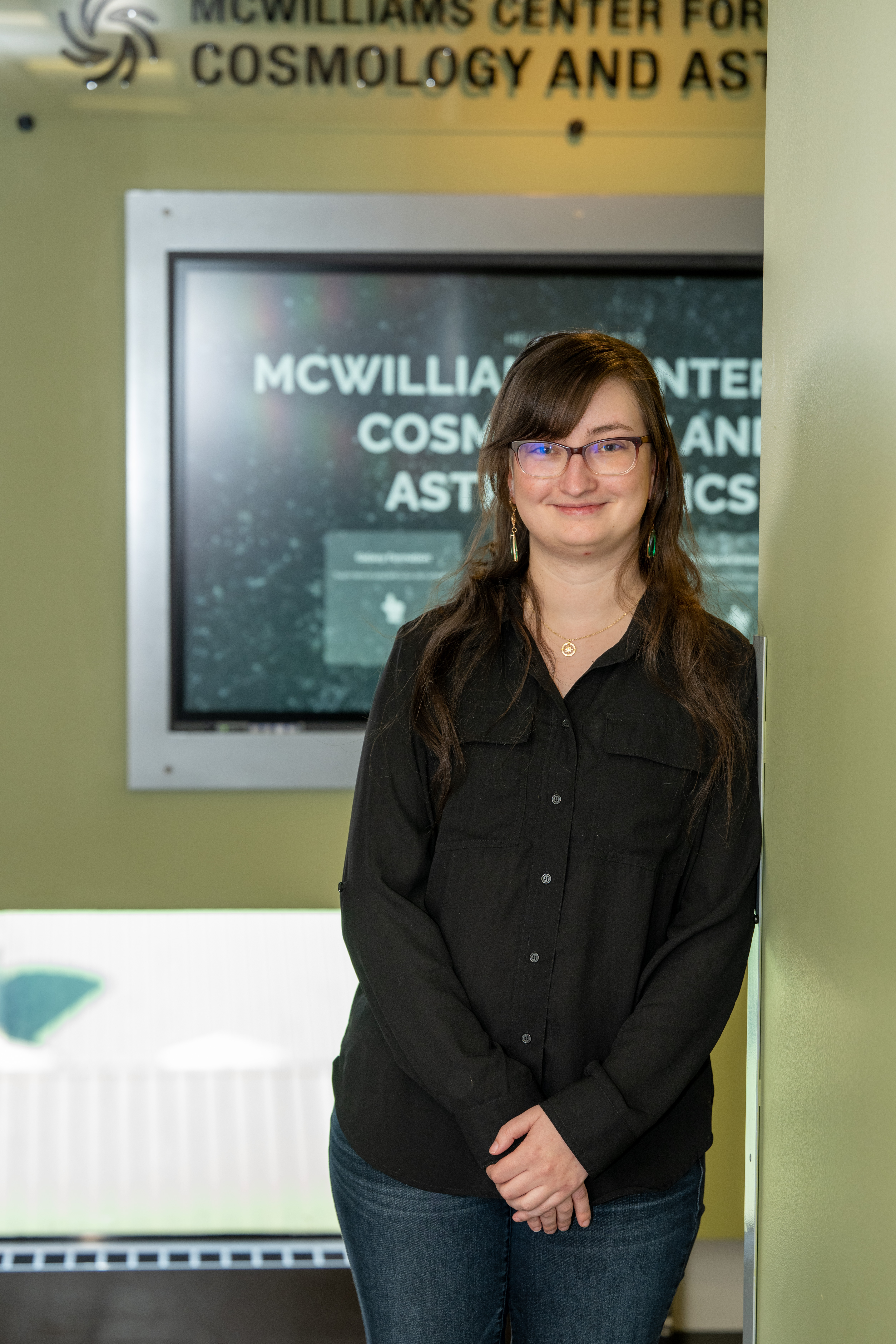 Anna O'Grady stands in front of a screen that advertises the McWilliams Center for Cosmology and Astrophysics. She leans against a bright green wall and smiles at the camera.