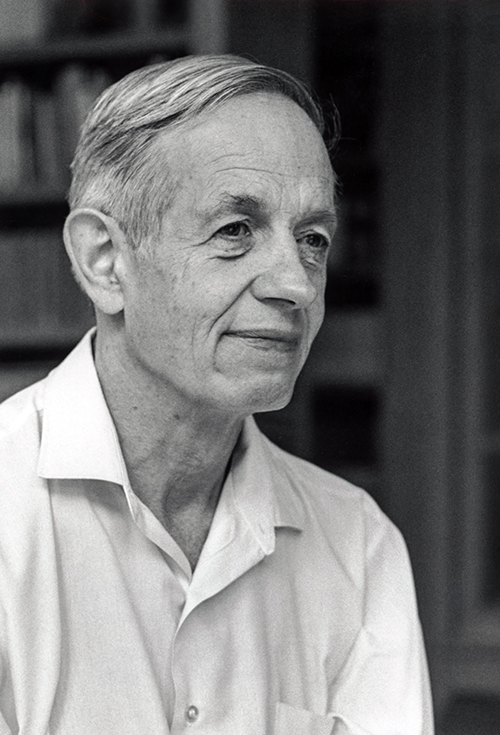 An older John Nash stands in front of the bookshelf and gazes into the distance. The photo is in black and white.