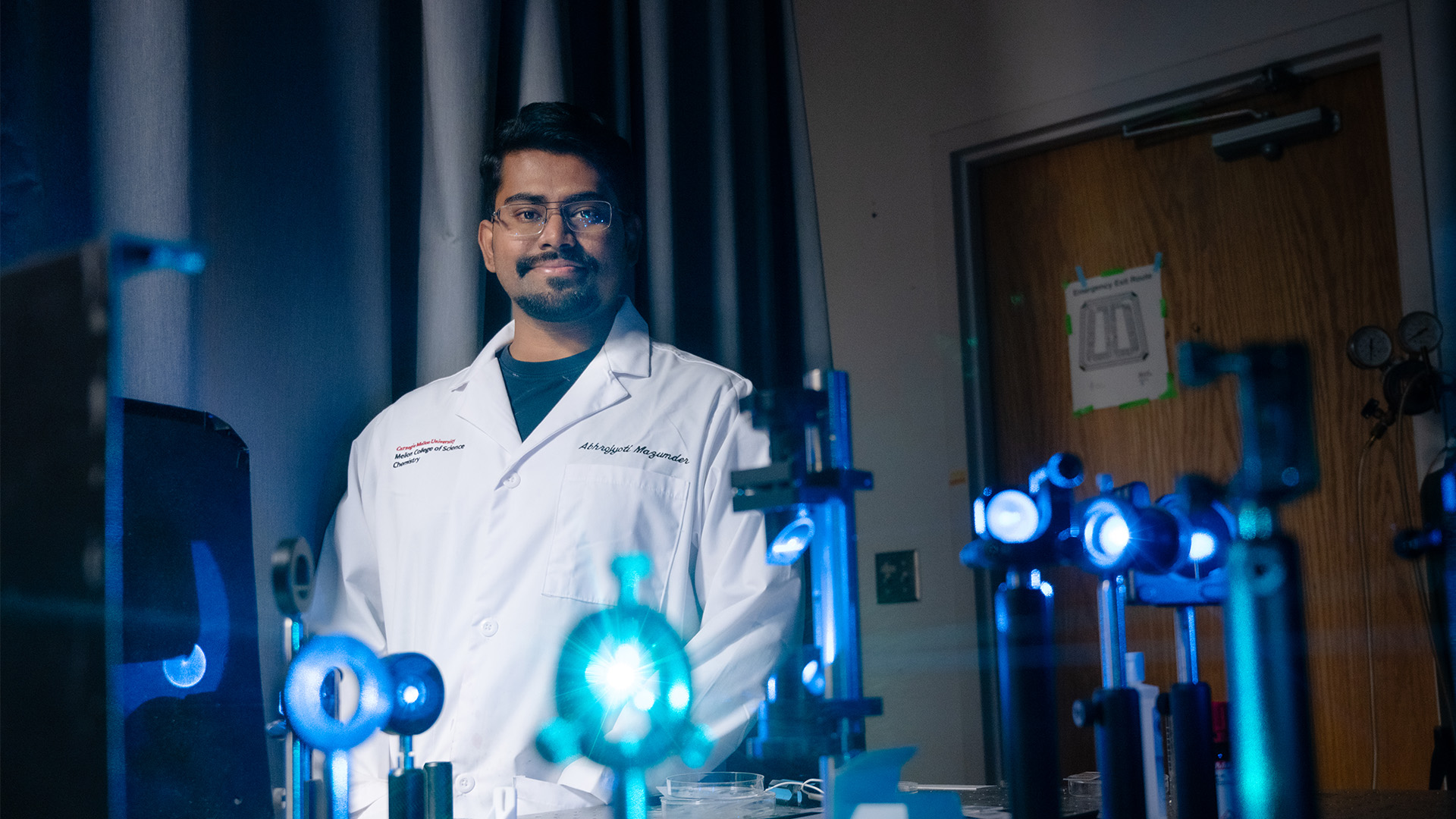 Abhrojyoti Mazumder stands in a lab coat. In front of him is a range of lenses with a blue laser passing through them. He smiles at the camera.