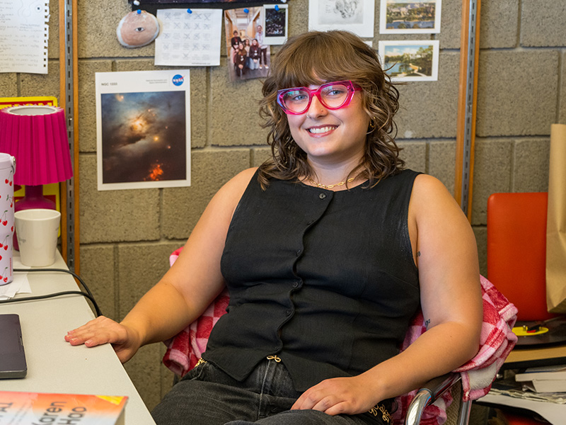 Sofia Splawska sits in her office and smiles at the camera. She is surrounded by photographs and books related to astrophysics and cosmology.
