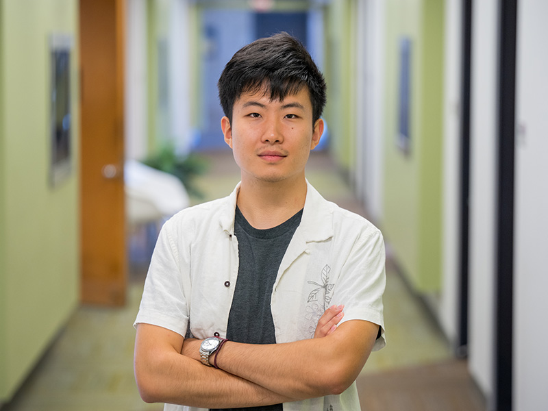 Eason Wang stands in a green hallway with brown wooden doors. He looks into the camera and crosses his arms.