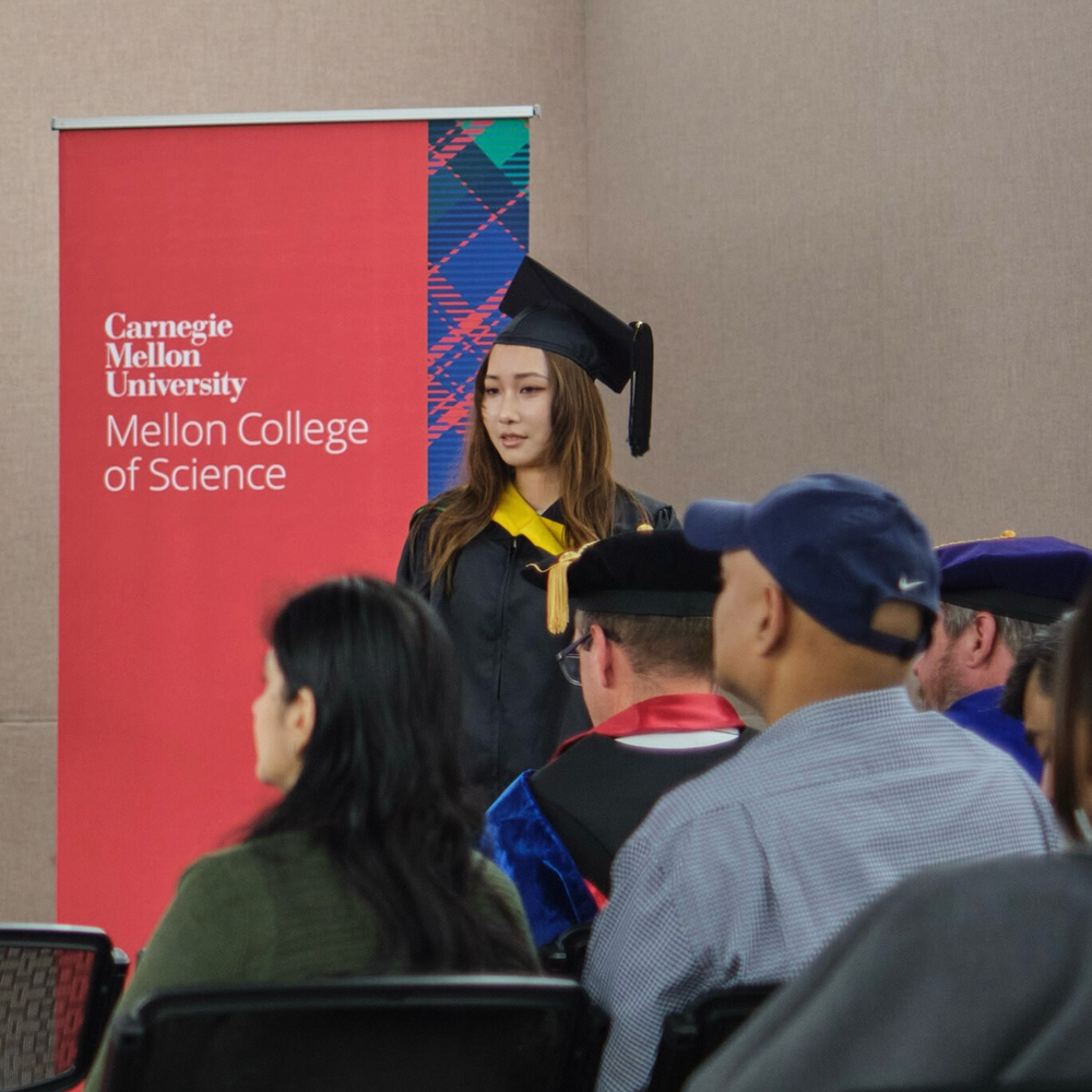 Rachel Tang stands in front of a room of people. She is wearing graduation regalia. She is next to a red and tartan sign that says Carnegie Mellon University Mellon College of Science.