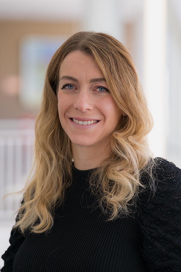 Antonella Palmese, a woman with long blonde hair, smiles warmly at the camera while standing in front of a window. She wears a black sweater.