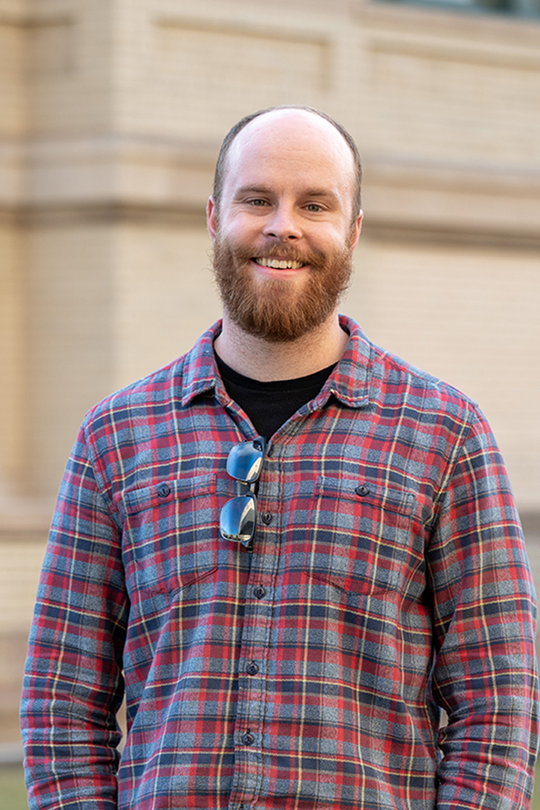 Smiling young man with a beard wearing a plaid shirt standing outdoors on campus.