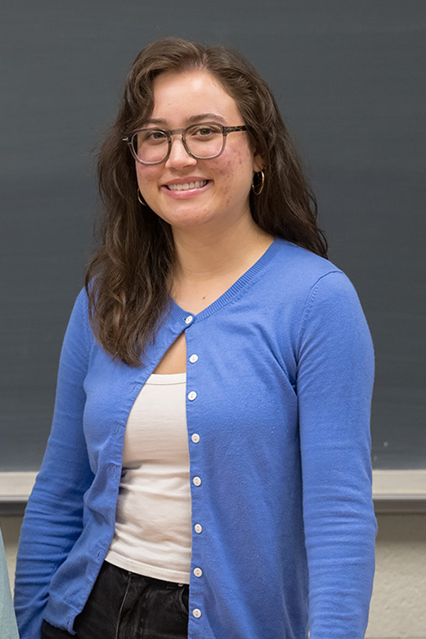 Smiling young woman wearing a blue cardigan standing in front of a blackboard.