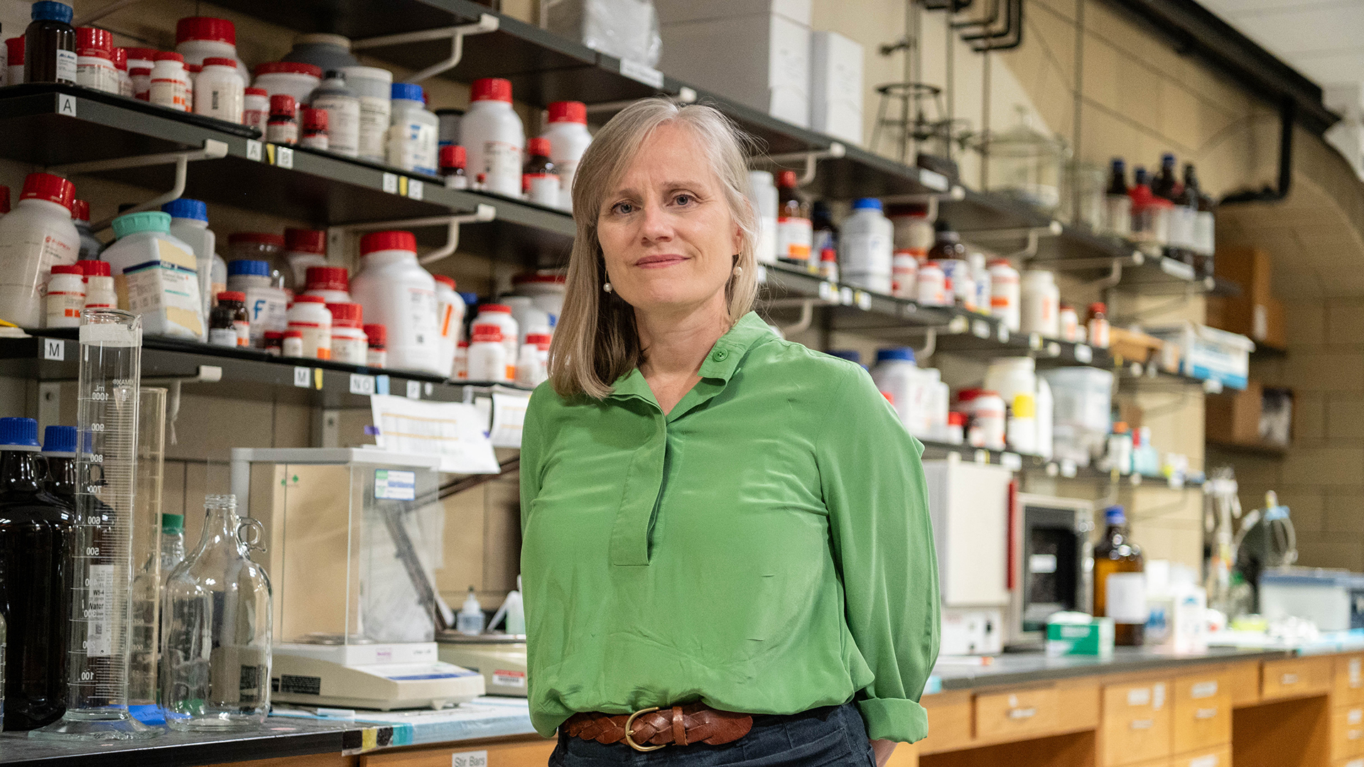 Alison Barth stands in her lab in front of a lab bench full of equipment and shelves full of bottles. She smiles at the camera.