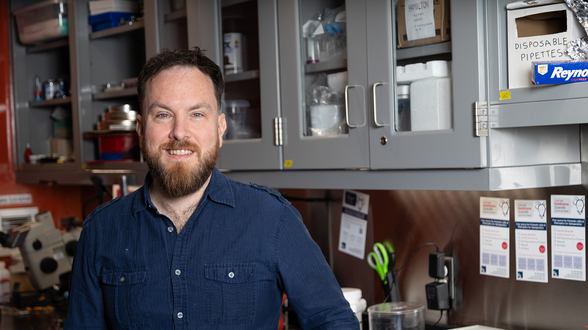 Eric Yttrium stands in his lab in front of cabinets full of equipment and a lab bench. He smiles at the camera.