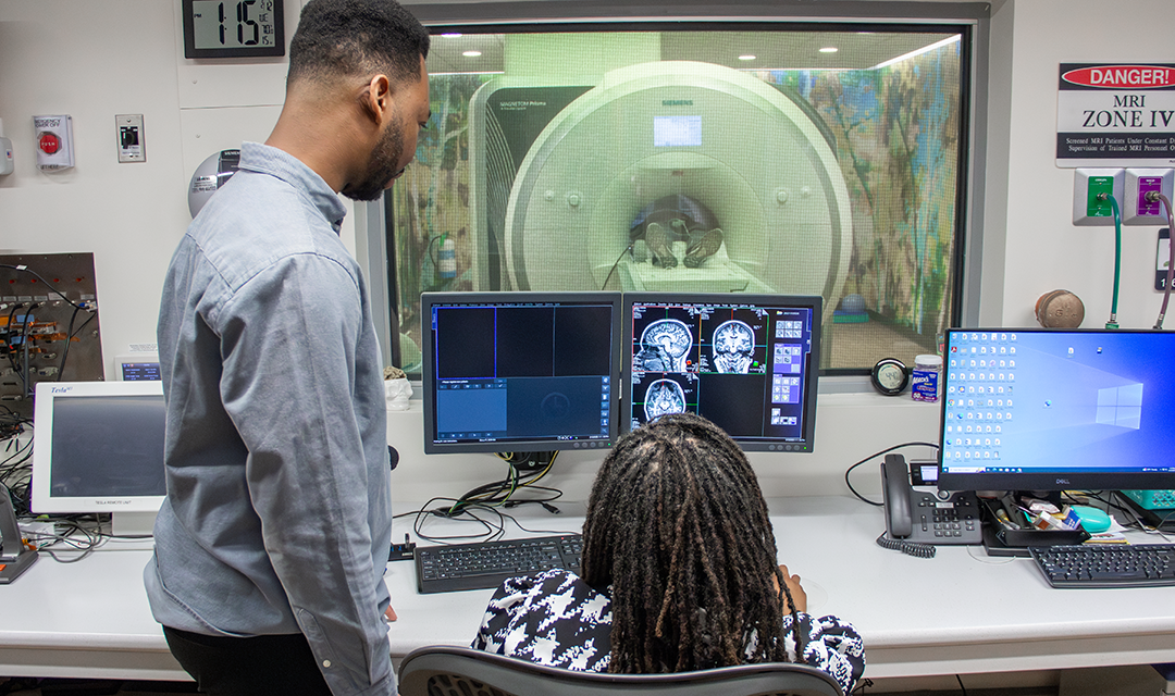 Two people watch an MRI happen and examine the images. In the background, a person lays in an MRI machine.