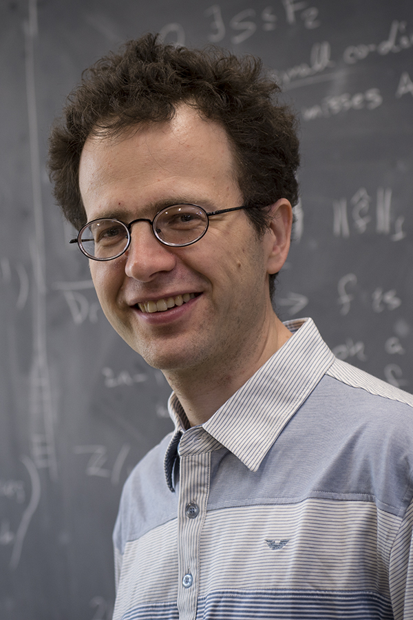 Boris Bukh smiles at the camera while standing in front of a chalkboard with math equations write on it.