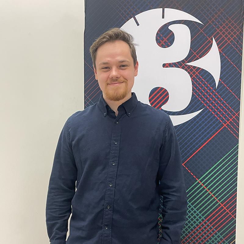 Christopher Blum stands in front of a Three Minute Thesis banner and smiles at the camera.