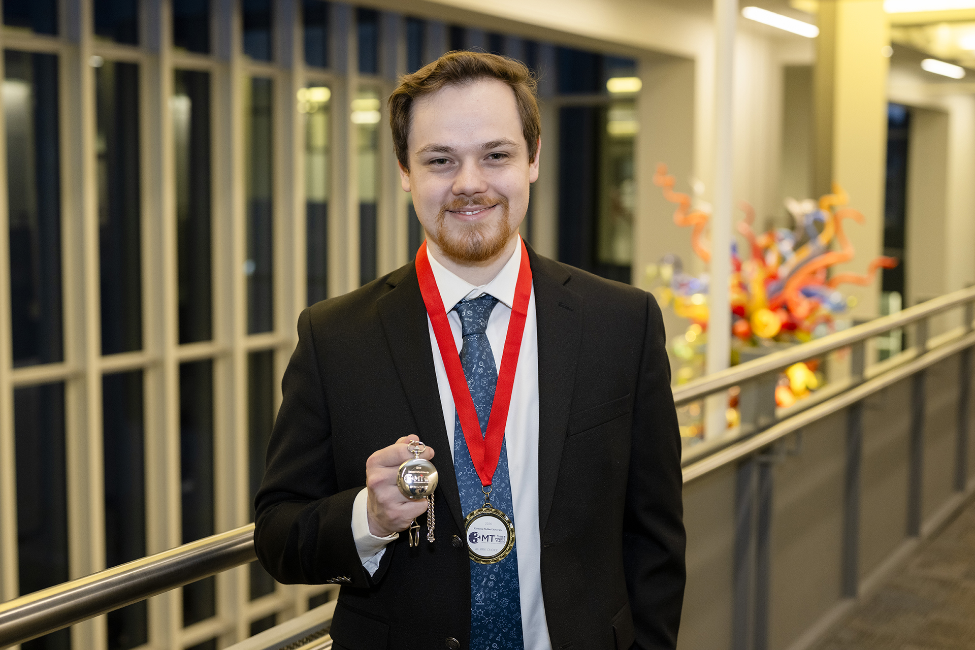 Christopher Blum stands in a hallway with a Three Minute Thesis medal and watch.
