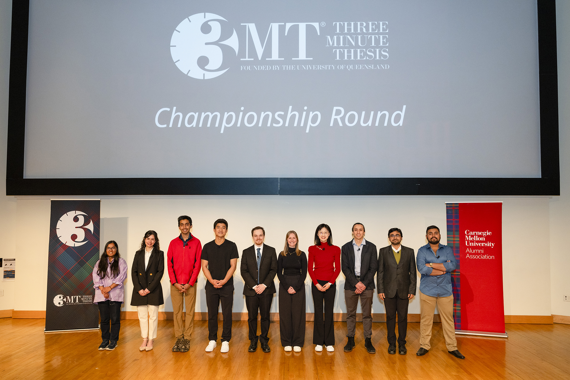 Ten Three Minute Thesis finalists stand on a stage in a line. They all look into the camera.
