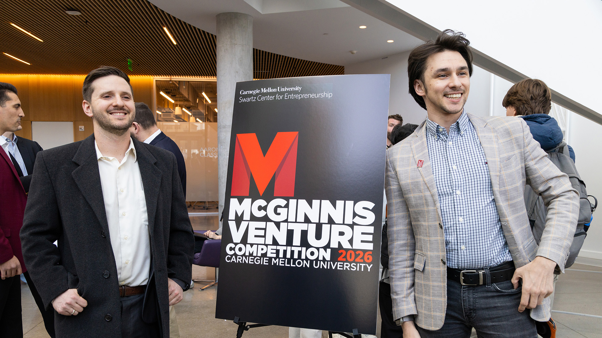 Ben Koby, wearing a black suit, and Filipp Gusev, wearing a plaid suit jacket and blue shirt, stand next to a McGinnis Venture Competition sign. Both men are smiling.