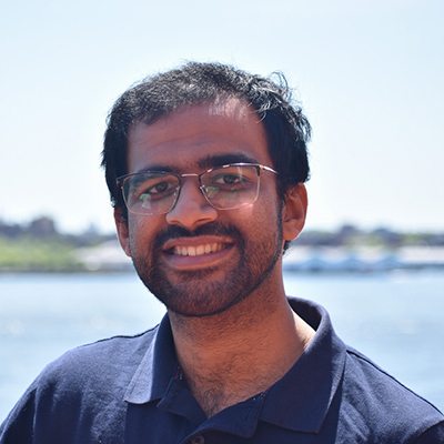 Pratyush Madapuji Ravi stands outside by a body of water. He smiles at the camera.