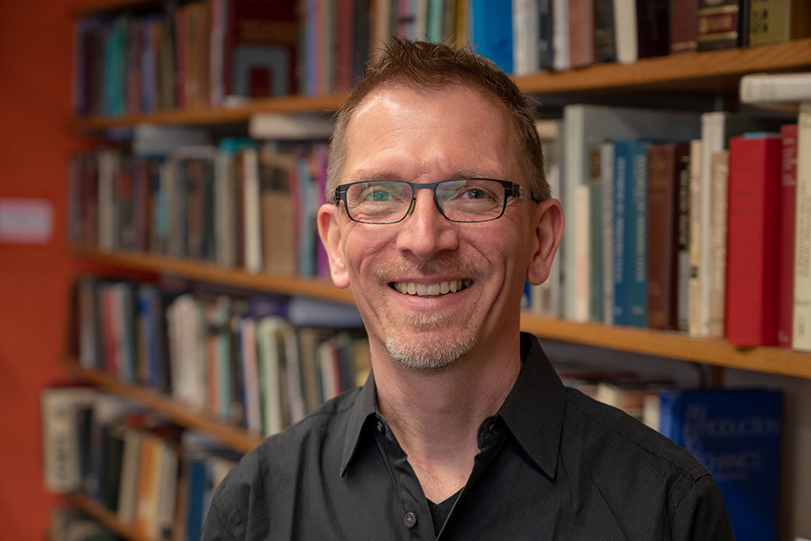 Markus Deserno, wearing glasses and smiling at the camera, stands in front of a bookcase full of colorful books.