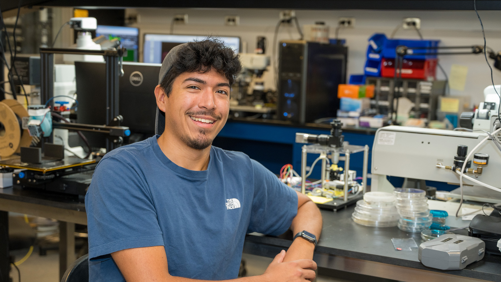 Brendan Ray sits in a physics lab. He smiles at the camera.
