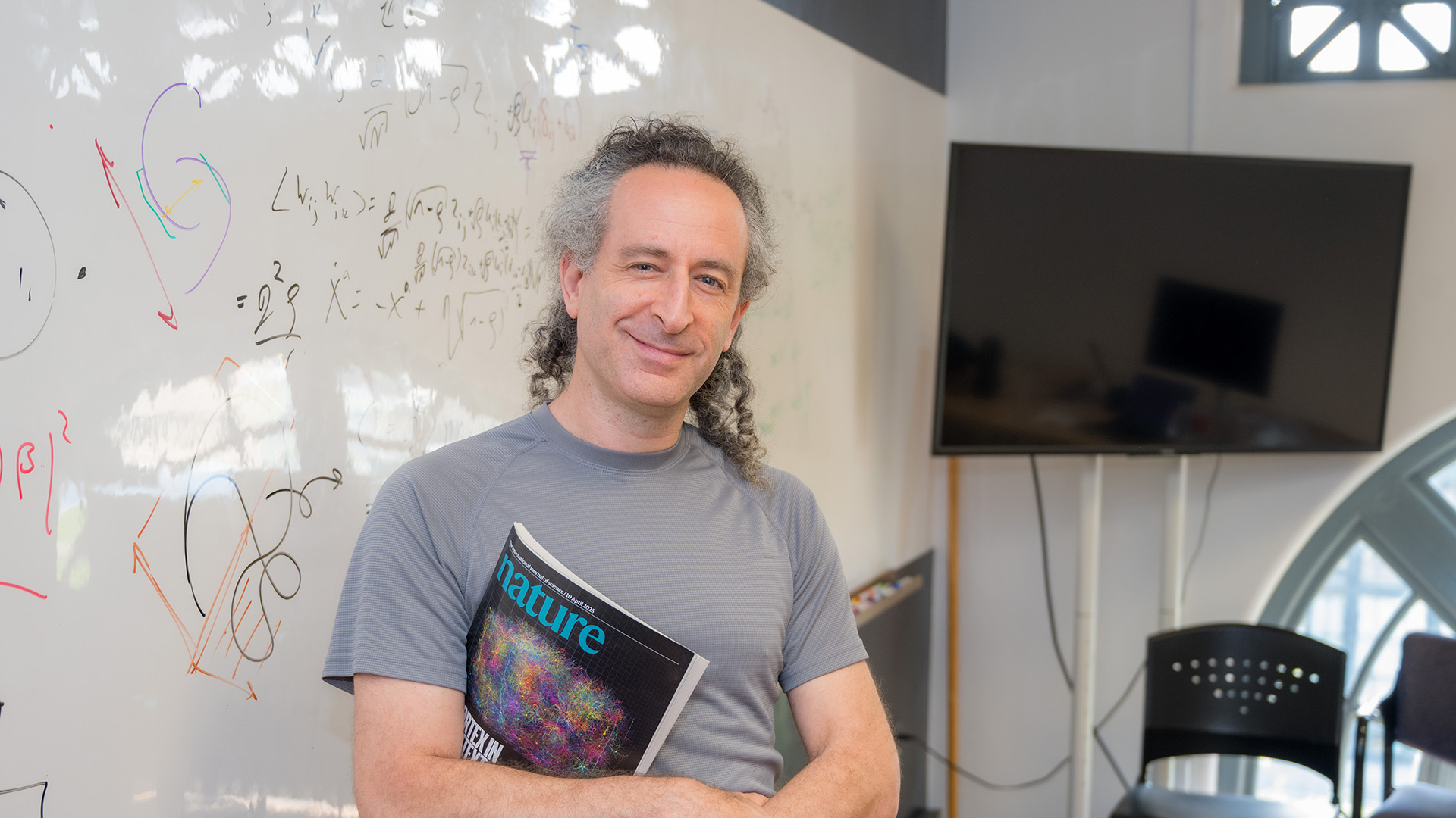 Xaq Pitkow stands in front of a whiteboard with writing on it. He holds a copy of Nature dedicated to the MICrONS project, and he smiles at the camera.