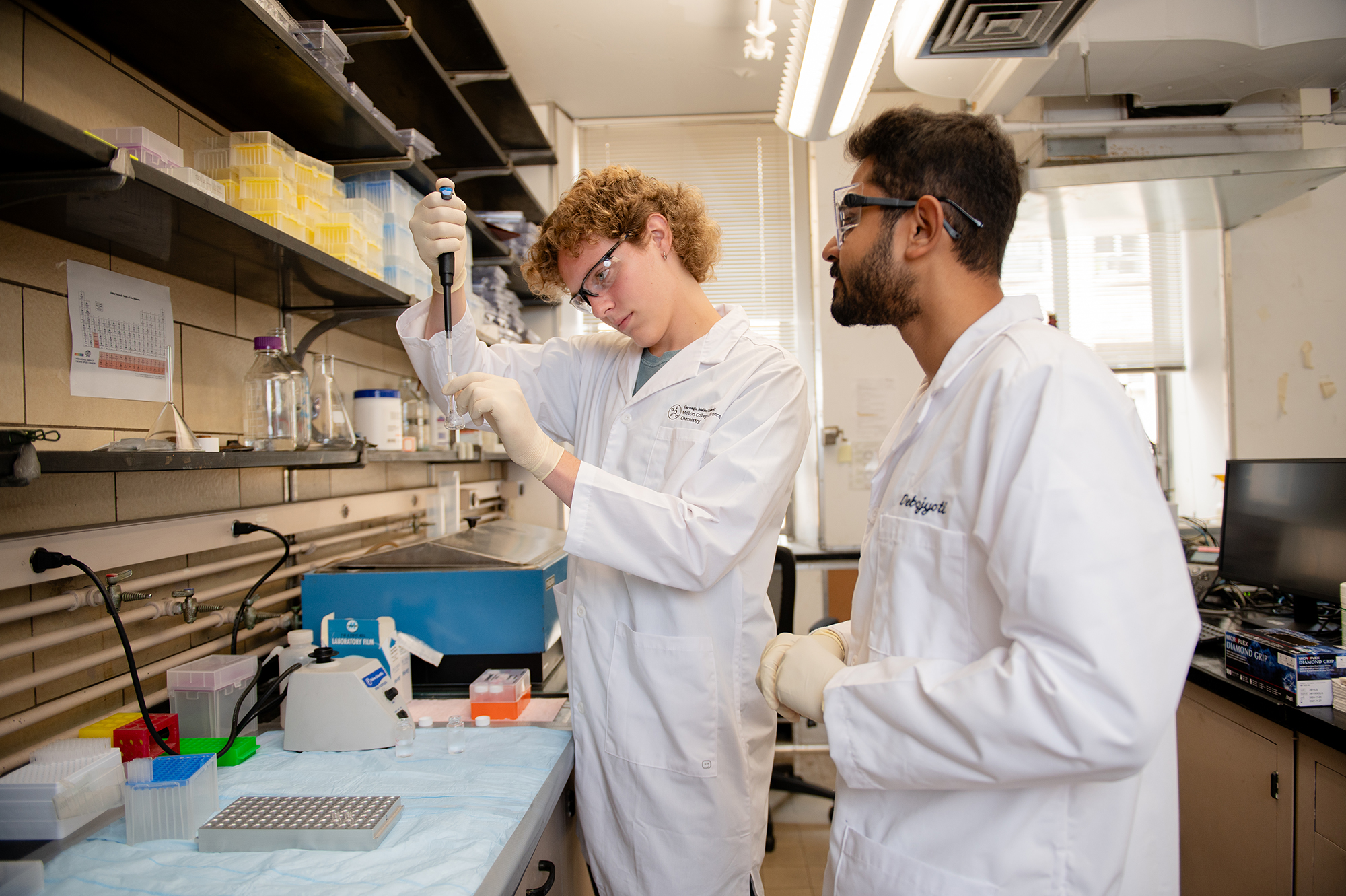 Matthew McCowen and Debojyoti Chakraborty stand at a lab bench. McCowen pipettes liquid into a test tube as Chakraborty watches.