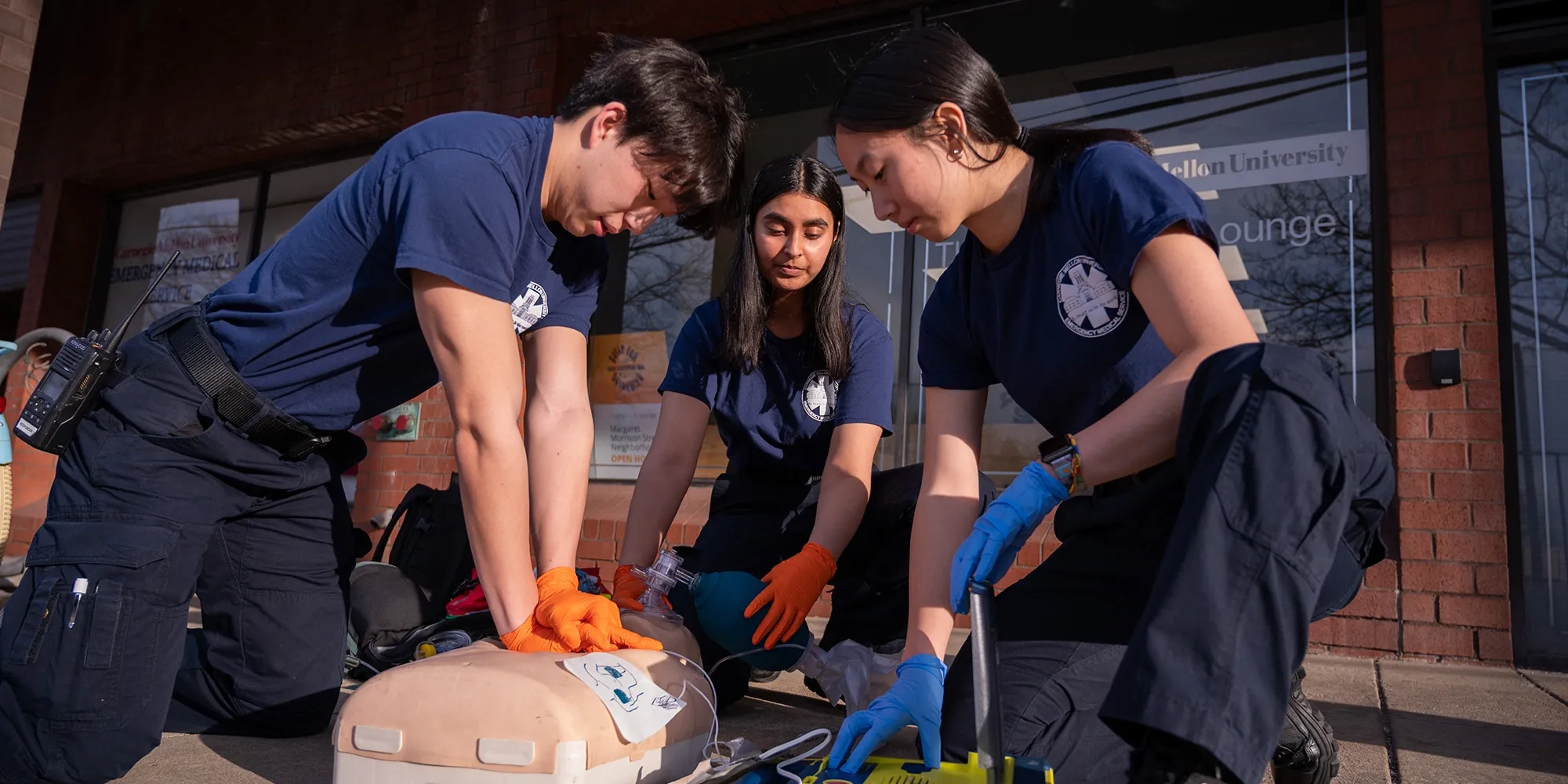 Three students—one male and two females—kneeling side by side, actively performing CPR on a training mannequin during a first aid class.