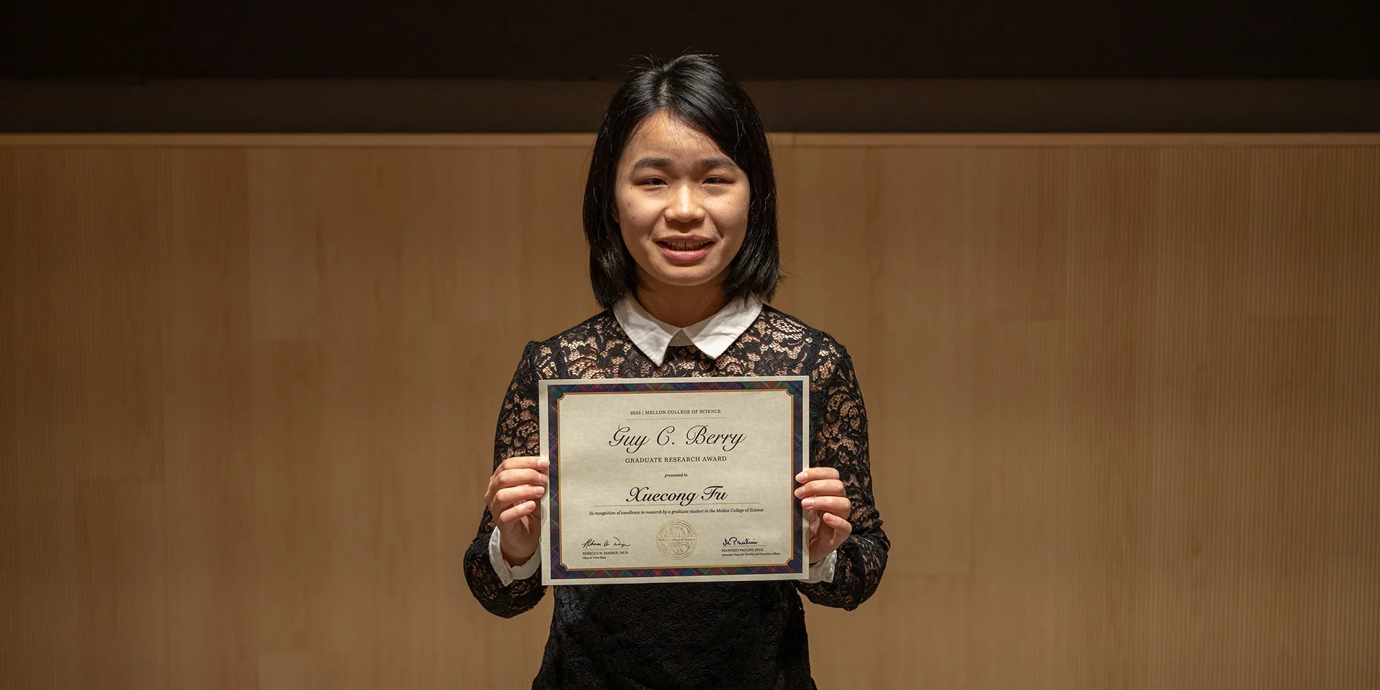 Young woman wearing a black lace top, smiling as she holds her diploma proudly in front of her.
