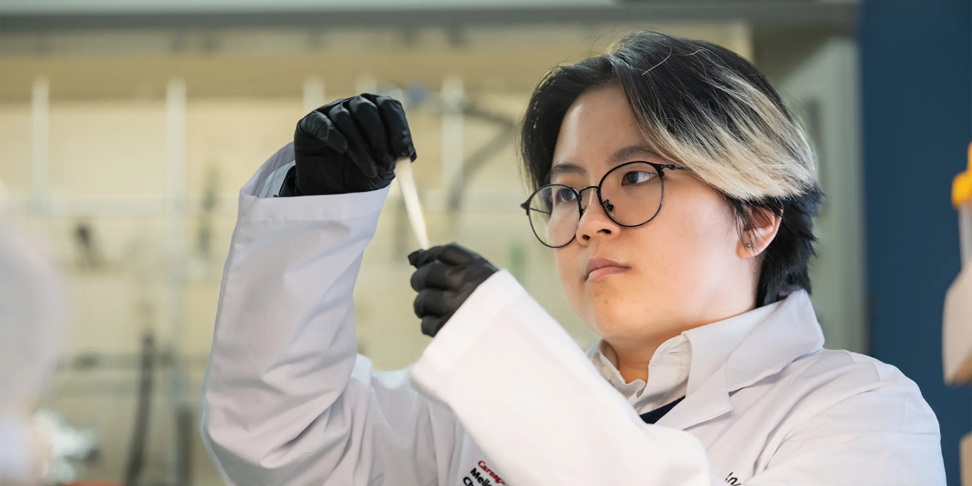 Tina Lin stands in a lab and stretches a polymer she created. She is wearing a lab coat, nitrile gloves and glasses.