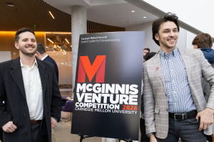 Ben Koby, wearing a black suit, and Filipp Gusev, wearing a plaid suit jacket and blue shirt, stand next to a McGinnis Venture Competition sign. Both men are smiling.