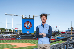 Calvin Hoffman stands in Kauffman Stadium surrounded by empty seats with the baseball diamond visible behind him.