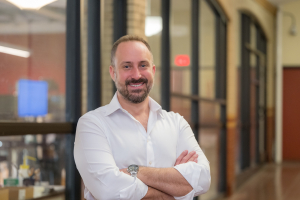 Smiling Nicholas Yoder in a white shirt stands in a hallway with his arms crossed, facing the camera.